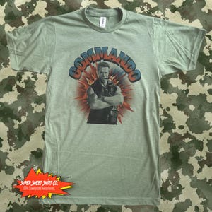 May include: Green t-shirt with a vintage-style graphic of Arnold Schwarzenegger from the movie Commando. The graphic features a red, orange, and yellow explosion behind Schwarzenegger, who is wearing a black tank top and holding a gun. The text "Commando" is in a blue and red font above Schwarzenegger's head.