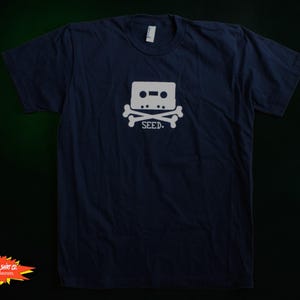 Puede incluir: Camiseta azul marino con un diseño gráfico blanco de una calavera con huesos cruzados en forma de cinta de cassette y la palabra "SEED" debajo. La camiseta es de material suave.