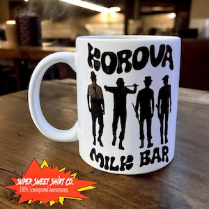 May include: White ceramic mug with a black graphic of four figures in silhouette with the text "KOROVA MILK BAR" printed on the front.