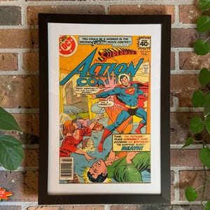 May include: A framed Action Comics Superman comic book cover featuring Superman in a blue and red suit, a woman in a red dress, and a man in a green shirt. The text reads Superman Action Comics, Great Krypton, and The Surprise Guest: Death.