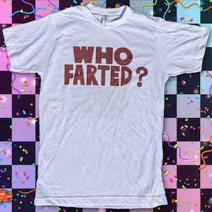 May include: A white short-sleeved t-shirt featuring the text "WHO FARTED?" printed in large, bold, dark brown capital letters across the chest. The garment has a standard crew neckline and a straight hemline, presented in a flat, front-facing view.