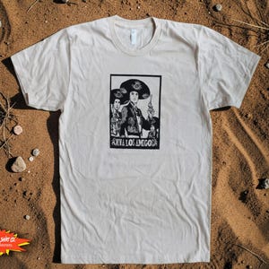 May include: Cream-colored t-shirt featuring a black and white graphic of two figures in traditional attire, one holding a gun. The text "VIVA LOS AMIGOS" is below the image. The shirt is laid on a sandy surface.