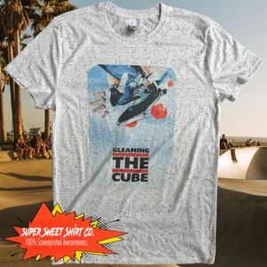 Può includere: Una maglietta grigia con un motivo a macchie bianche. La maglietta presenta una grafica di uno skater che esegue un trick con il testo "GLEAMING THE CUBE" sotto. La maglietta ha anche un logo rosso, giallo e bianco che dice "SUPER SWEET SHIRT CO. 100% Screenprinted Awesomeness."