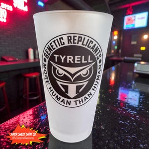 May include: A frosted glass with a black and white graphic. The graphic features the text "GENETIC REPLICANTS TYRELL" around an owl-like design. The text "MORE HUMAN THAN HUMAN" is also present. The bottom left corner has the text "SUPER SWEET SHIRT CO."