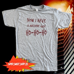 May include: A gray t-shirt with the text "NOW I HAVE A MACHINE GUN HO-HO-HO" printed in maroon. The text is in a bold, comic-book style font. The shirt is from Super Sweet Shirt Co.