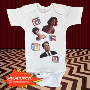 May include: A white baby grow with a red, white and blue colour scheme. The grow features images of characters from the TV show Twin Peaks, including Agent Cooper, Audrey Horne, and a young girl. The grow also has the letters "TWIN" spelled out with blocks.