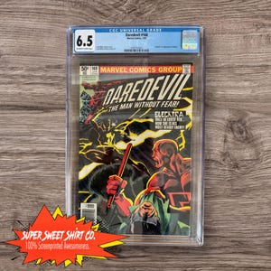 May include: A graded comic book in a plastic case. The comic book cover features a red and black superhero character, Daredevil, fighting a female character, Elektra. The comic book is titled "Daredevil" and the cover text reads "The Man Without Fear!" and "Elektra: Once he loved her... Now she is his most deadly enemy!"