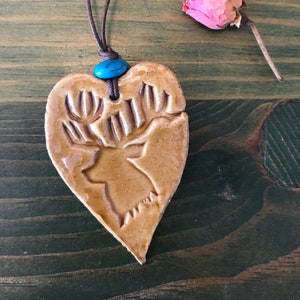 Puede incluir: Collar con colgante en forma de corazón con diseño de ciervo. El colgante es de color beige con una cabeza de ciervo y cornamenta talladas. Una pequeña cuenta turquesa está en la parte superior del colgante, y un cordón marrón forma el collar. Una rosa rosa seca está al lado.