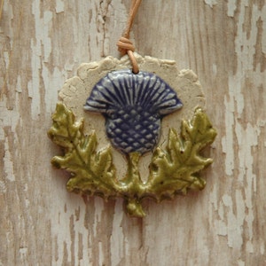 May include: A ceramic pendant necklace featuring a purple thistle with green leaves. The pendant is on a brown leather cord and is hanging against a white wooden background.
