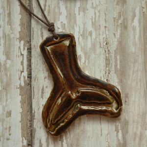 Könnte beinhalten: Ein brauner Keramik-Anhänger in Form eines Fußes mit weißem Überzug. Der Anhänger hängt an einer braunen Lederschnur.
