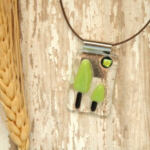 Puede incluir: Un collar con un colgante de vidrio verde con dos árboles y una pequeña piedra preciosa verde. El collar está en un cordón marrón y está colgando sobre un fondo de madera marrón claro.