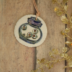 May include: A white ceramic ornament with a blue and purple design of a jar with a lid. The ornament has a brown cord for hanging.
