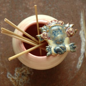 Może przedstawiać: Ceramiczny dyfuzor z różowym szkliwem i ozdobnym ceramicznym elementem wierzchnim z kwiatowym wzorem. Dyfuzor zawiera drewniane patyczki.