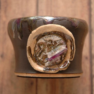 May include: A dark brown ceramic mug with a light brown glaze. The mug has a circular design with a silver and iridescent crystal embedded in the center.