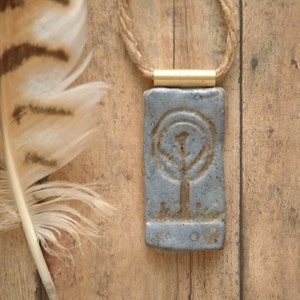 Puede incluir: Un colgante de cerámica azul con un diseño de árbol cuelga de un collar de cordón marrón. El colgante es rectangular y tiene una textura rugosa. El collar está sobre una superficie de madera.
