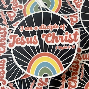 May include: A round sticker with the text "I am a disciple of Jesus Christ" in a retro font. The design features a black background with white radial lines and a rainbow. The text "3 Nephi 5:13" is also included.