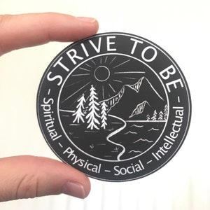 May include: Black and white sticker with the text "Strive to Be" in a circle. The text is surrounded by a mountain scene with a sun, trees, and a river. The words "Spiritual", "Physical", "Social", and "Intellectual" are written around the circle.