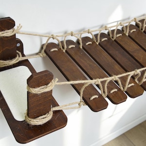 Puede incluir: Un puente para gatos de madera marr&oacute;n oscuro, con tablones de madera unidos por cuerda natural. El puente incluye una plataforma rectangular blanca y est&aacute; dise&ntilde;ado para montaje en pared.