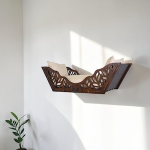 Puede incluir: Una cama para gatos montada en la pared, hecha de madera marr&oacute;n oscuro, con un dise&ntilde;o de hojas y el nombre "Felix" cortado en la parte delantera. Tiene un coj&iacute;n de color crema en el interior. La cama est&aacute; montada en una pared blanca.