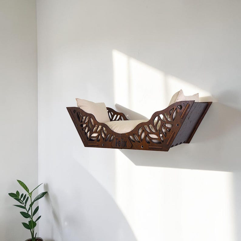 Puede incluir: Una cama para gatos montada en la pared, hecha de madera marr&oacute;n oscuro, con un dise&ntilde;o de hojas y el nombre "Felix" cortado en la parte delantera. Tiene un coj&iacute;n de color crema en el interior. La cama est&aacute; montada en una pared blanca.