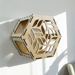 Puede incluir: Una estanter&iacute;a de pared hexagonal de madera clara con un dise&ntilde;o recortado con forma de hojas. La estanter&iacute;a est&aacute; montada en una pared blanca, mostrando su forma geom&eacute;trica y elementos decorativos. La estanter&iacute;a tiene dos compartimentos.