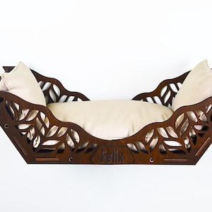 Puede incluir: Una cama para gatos de madera marrón oscuro, con recortes en forma de hojas y un cojín color crema. La cama tiene un diseño curvo y el nombre "Felix" grabado en la parte delantera.