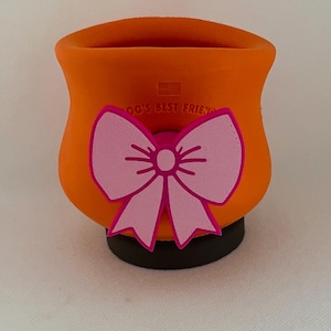 Op de afbeelding: Een oranje dierenvoedseldispenser met een roze strik. De dispenser heeft de tekst "DOG'S BEST FRIEND" en een kleine Amerikaanse vlag. De dispenser staat op een zwarte basis.