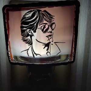 May include: A night light featuring a black silhouette of a person wearing sunglasses. The image is set against a pinkish background within a rectangular frame. The night light is plugged into an electrical outlet.