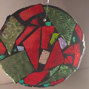 May include: A round, stained-glass ornament with a mosaic design. The ornament features red, green, and iridescent glass pieces set against a black background. A small hole is at the top for hanging. The ornament is suspended by a thin, white string.
