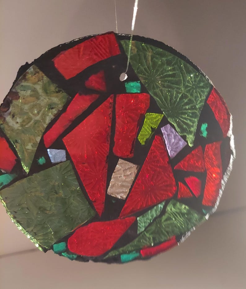 May include: A round, stained-glass ornament with a mosaic design. The ornament features red, green, and iridescent glass pieces set against a black background. A small hole is at the top for hanging. The ornament is suspended by a thin, white string.