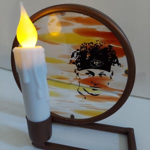 May include: A decorative candle holder featuring a battery-operated candle with a flickering yellow flame. The candle sits in front of a round, stained-glass-style panel with a brown frame. The panel has a silhouette of a person.
