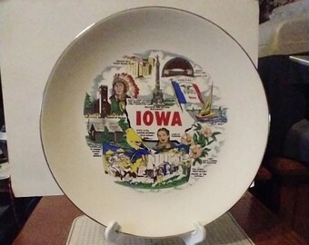 May include: A vintage souvenir plate featuring a colorful map of Iowa with landmarks and the word "IOWA" in red. The plate has a gold rim and is displayed on a white stand.
