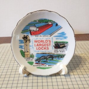 May include: A white ceramic souvenir plate with a gold rim. The plate features a colorful illustration of the Soo Locks in Sault Ste. Marie, Michigan. The text on the plate reads "World's Largest Locks".