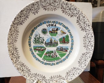 May include: A white ceramic plate with a blue and gold floral border. The center of the plate features a colorful illustration of the state of Iowa with the text "IOWA" and "THE HAWKEYE STATE".