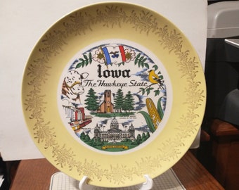 May include: A yellow ceramic plate with a gold rim and a colorful illustration of Iowa landmarks. The plate features the text "Iowa The Hawkeye State" and depicts the state capitol building, cornfields, and a bird.