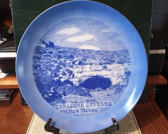 May include: Blue and white ceramic souvenir plate featuring a scenic image of Carlsbad Caverns, New Mexico. The plate has a gold rim and the text "Carlsbad Caverns New Mexico" printed on the bottom.