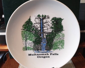 Puede incluir: Plato de cerámica blanco con una ilustración en negro y verde de Multnomah Falls en Oregón. El plato está sobre un soporte negro.