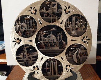 May include: A silver-colored metal plate with a black and copper design featuring various landmarks of Istanbul, Turkey. The plate is on a stand and has a textured surface.
