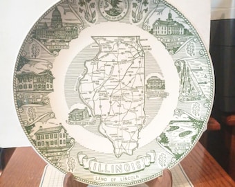 May include: A vintage souvenir plate featuring a map of Illinois in the center, surrounded by green illustrations of landmarks and the state seal. The plate is displayed on a wooden stand, showcasing the state's name and the phrase "Land of Lincoln."