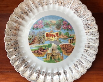 May include: A white ceramic plate with a gold rim and a colorful illustration of Iowa landmarks. The plate features the text "Iowa Hawkeye State" and images of the State Capitol, a field of corn, and a scene of Native Americans.