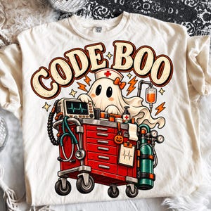 Puede incluir: Una camiseta color hueso con un gráfico de un fantasma con cofia de enfermero detrás de un carro médico rojo. El carro incluye un monitor cardíaco, estetoscopio, bolsa de suero y tanque de oxígeno. El texto CODE BOO está impreso en letras grandes y negritas sobre el fantasma.