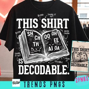 Op de afbeelding: Een zwart t-shirt met korte mouwen en een witte afbeelding van een open boek met fonetische tekst zoals SH, CH, TH, OO, OH, EE, AI en OA. Het shirt toont de tekst THIS SHIRT IS DECODABLE met pijlen die wijzen naar termen als BLEND, SOUND IT OUT, WORD CHUNK, SYLLABLE en READ IT.