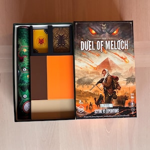 Könnte beinhalten: Eine Brettspielschachtel mit dem Titel Duel of Meloch, die eine Fantasy-Illustration eines Reisenden mit Gewehr und Bär zeigt. Die offene Schachtel enthält eine aufgerollte grüne Karte, Kartenstapel mit gelben und braunen Rückseiten sowie leere orangefarbene und beige Ablagefächer.