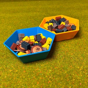 以下が含まれることがあります： 様々なゲームコンポーネントが入った、青とオレンジの2つの六角形のゲームピースホルダー。青いホルダーには、黄色、茶色、グレー、丸いピースが入っています。オレンジ色のホルダーにも同様のピースが入っています。緑色のテクスチャのある表面に置かれています。