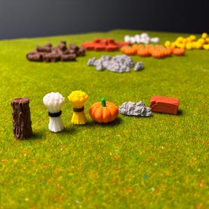 Op de afbeelding: Miniatuur boerderijscène met diverse elementen. Bevat een bruine boomstronk, witte en gele tarwebundels, een oranje pompoen, grijze rotsen en een rode baksteen. De items zijn gerangschikt op een groen oppervlak.
