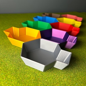 May include: A selection of brightly coloured, geometric trays in shades such as yellow, green, blue, red, purple, and grey. The trays are arranged on a green surface, highlighting their angular design and vibrant colours. These trays are ideal for organizing small items.