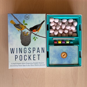 Puede incluir: Caja del juego de mesa Wingspan Pocket y bandeja abierta. La portada muestra dos oropéndolas y un nido con polluelos. La bandeja de plástico azul verdoso contiene huevos pequeños de color rosa moteado, un mazo de cartas y cinco cubos de madera.
