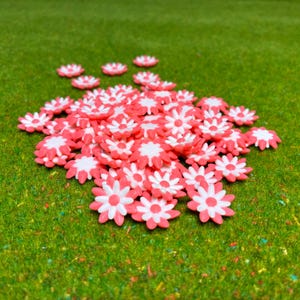 May include: A collection of small, pink and white daisy-shaped embellishments scattered on a green surface. The flowers have a white center and pink petals, creating a cheerful and delicate appearance. These decorative elements are ideal for craft projects.