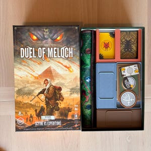 Könnte beinhalten: Eine Brettspielschachtel mit dem Titel Duel of Meloch und einer Fantasy-Illustration einer Person mit Gewehr und einem Bären in einer Wüstenlandschaft. Der offene Schachtelinhalt zeigt eine grüne Spielmatte, Kartenstapel und Spielmarken.