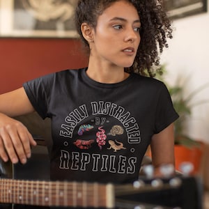 May include: Black t-shirt with the text "EASILY DISTRACTED BY REPTILES" in a circular design. The design includes illustrations of various reptiles in shades of red, purple, and brown. A black acoustic guitar is in the foreground.
